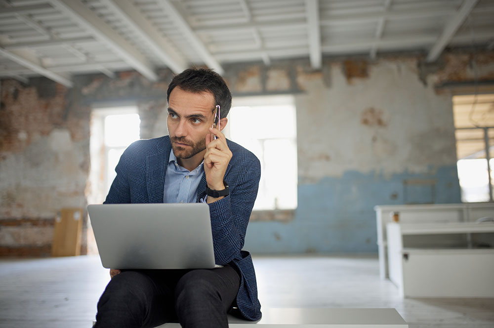 Portrait of man, businessman, worker sitting with laptop with thoughtful expression in big empty room. Thinking process, making analytics